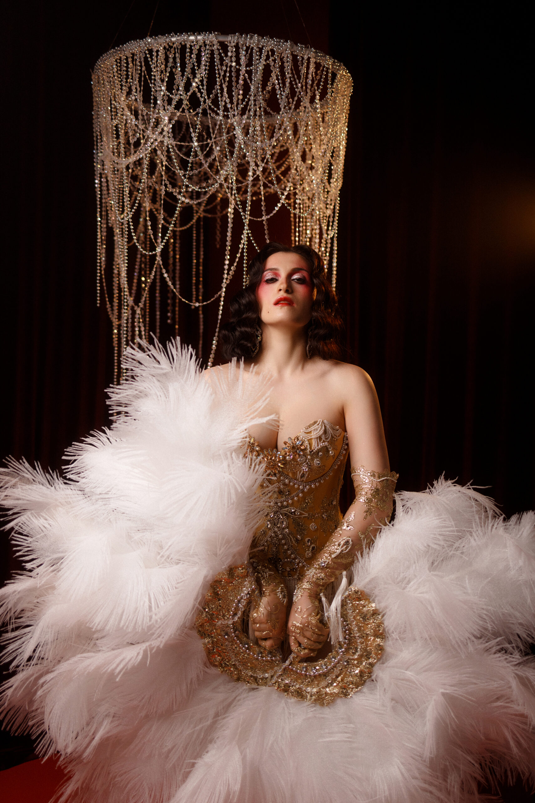 Young Caucasian circus performer or burlesque dancer in golden vintage corset, front view, posing under chandelier on dark background. Concepts: circus, cabaret, retro, show, performance.